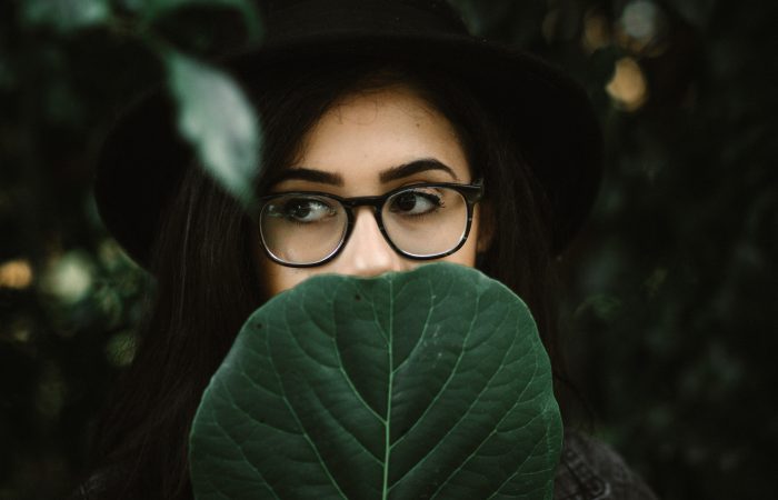 artistic photo of young woman with dark hair and glasses, a black hat, with a large dark geen leaf covering the lower half of her face