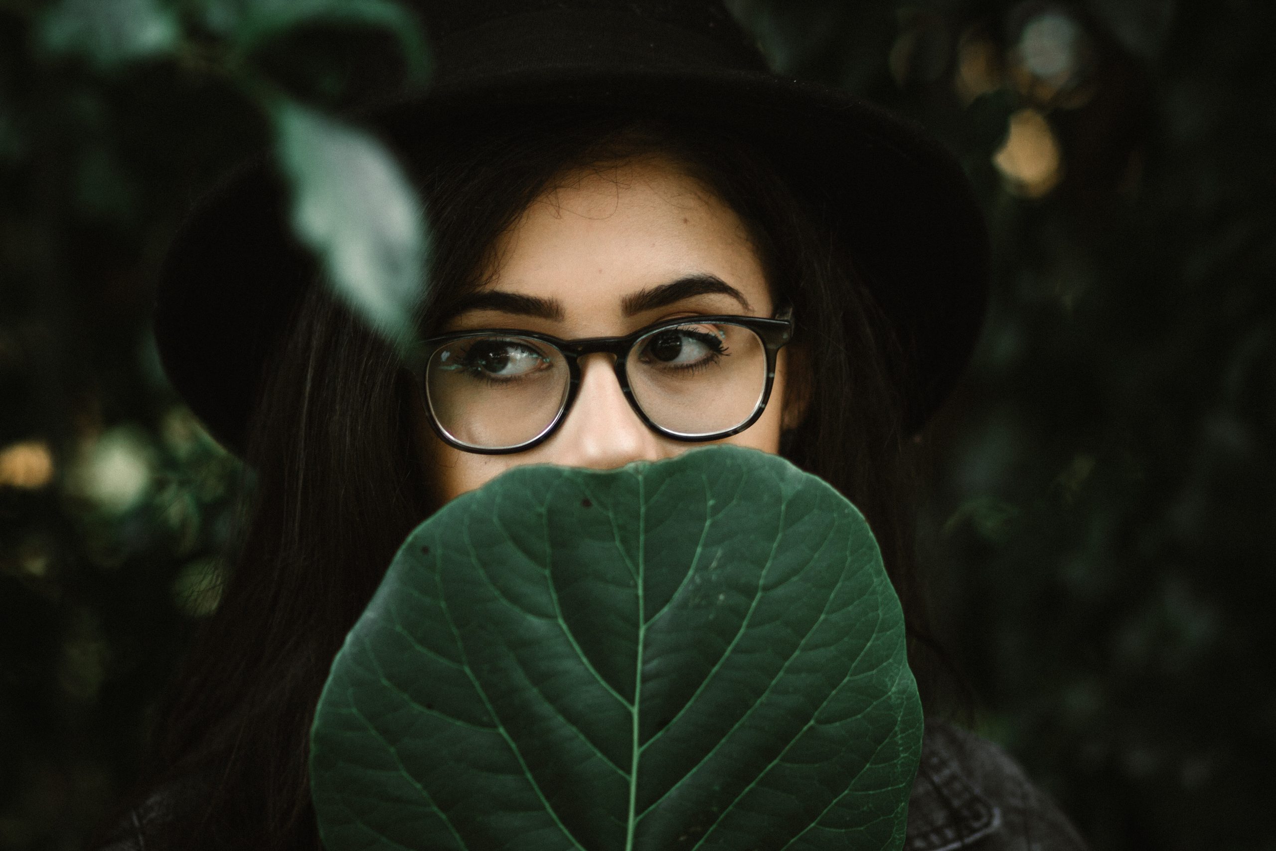 artistic photo of young woman with dark hair and glasses, a black hat, with a large dark geen leaf covering the lower half of her face