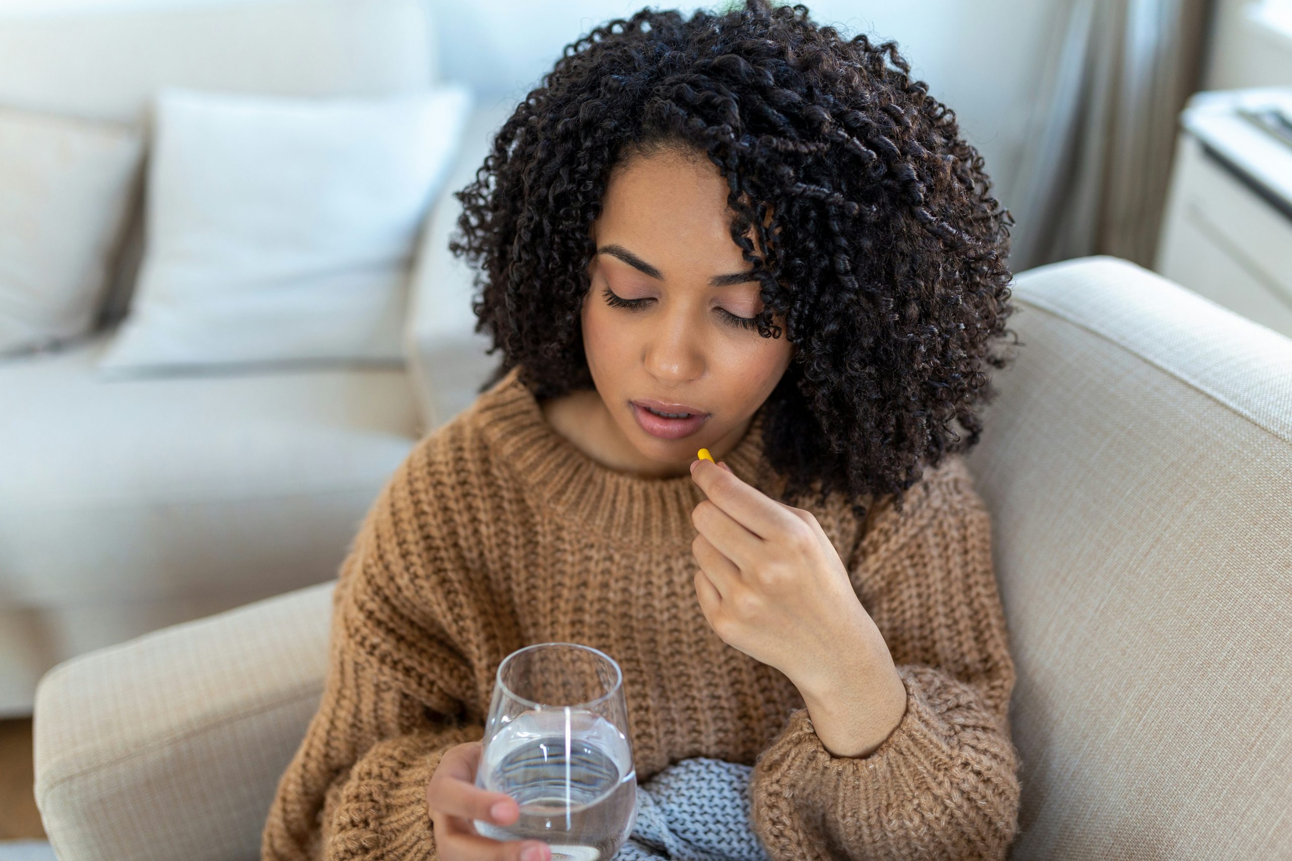 a young black woman sitting on a couch taking a pill with a glass of water